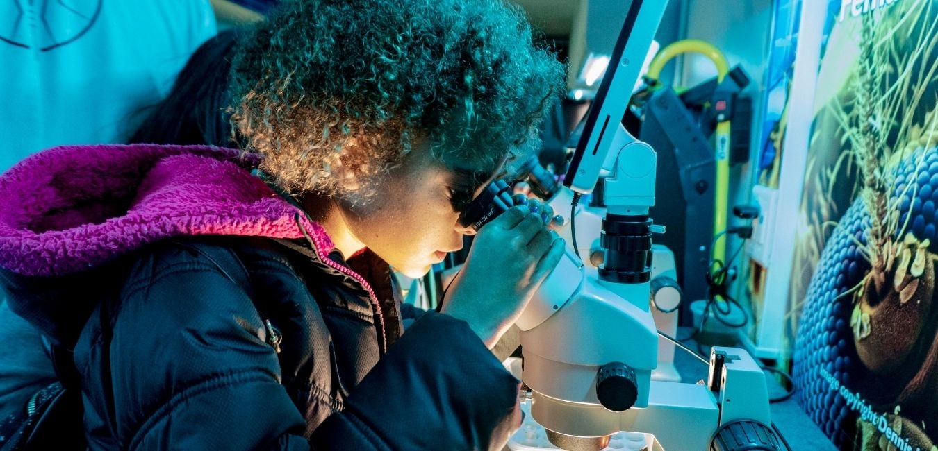 A child with curly hair looks through a microscope, engaged in exploration or learning, with colorful lighting in the background.
