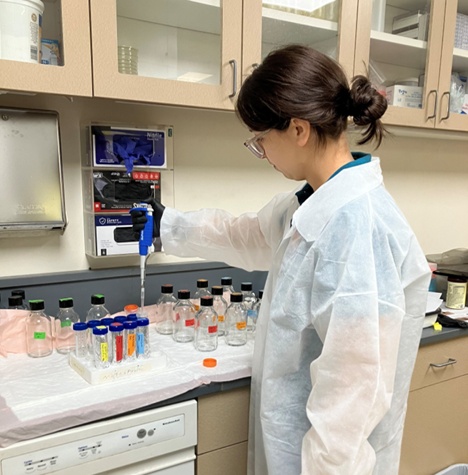 Person in a lab coat using a pipette with bottles on a covered bench in a laboratory setting.