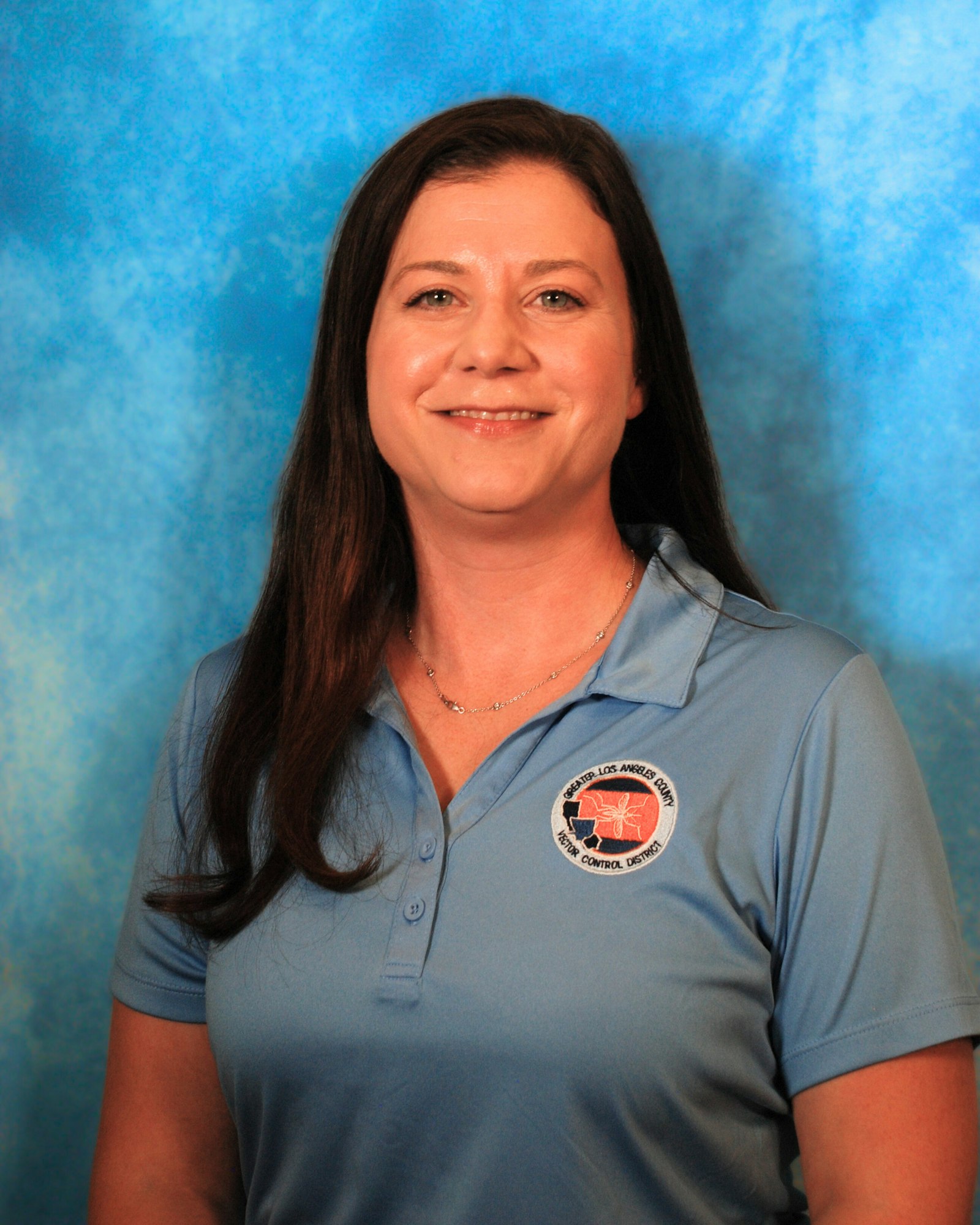 A person with long hair is wearing a blue polo shirt with a logo against a blue background.