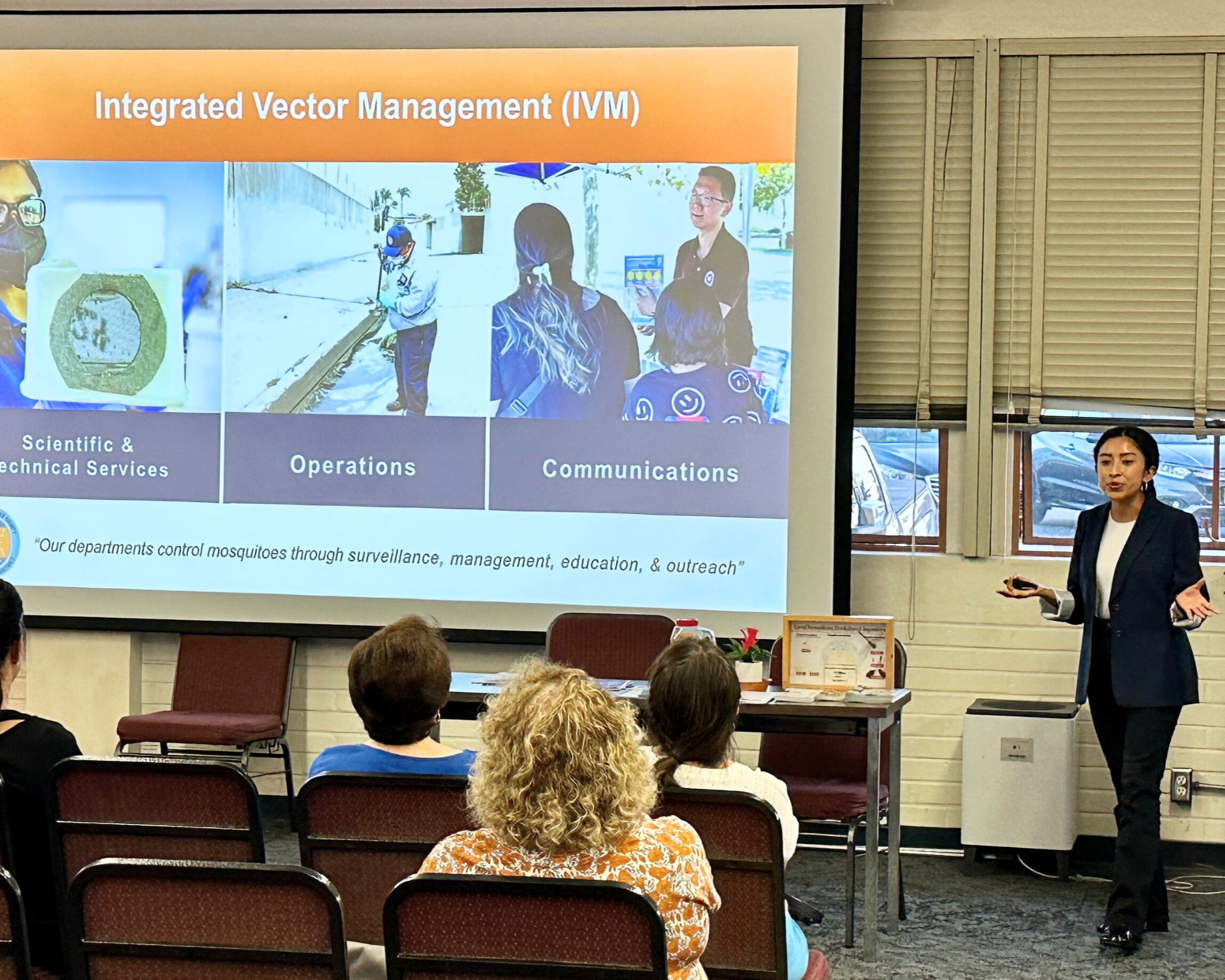 A woman presents on Integrated Vector Management (IVM) to an audience, covering scientific services, operations, and communications.