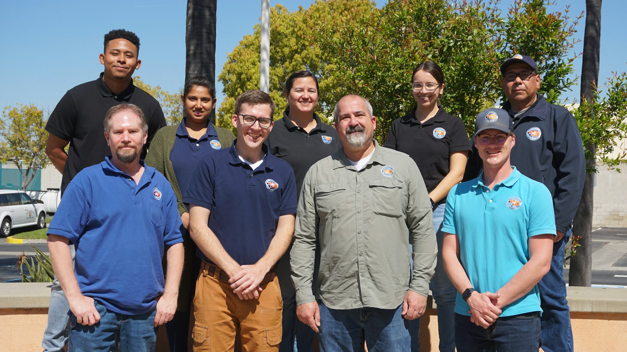 A group portrait of nine people, smiling, outdoors, wearing matching shirts, with trees and vehicles in the background.
