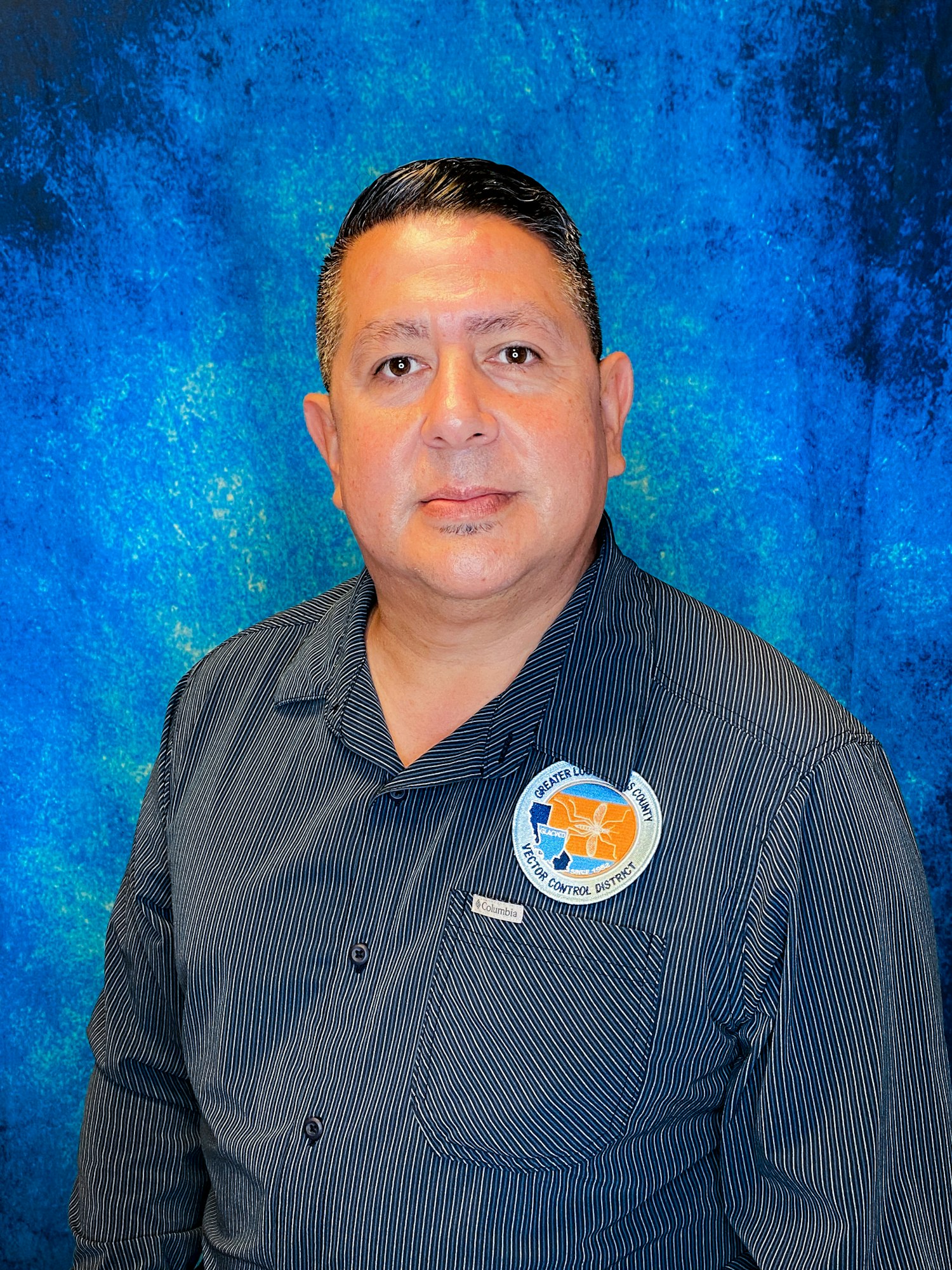 A person in a striped shirt with a "Greater Lodi" badge on the chest stands against a blue textured background.