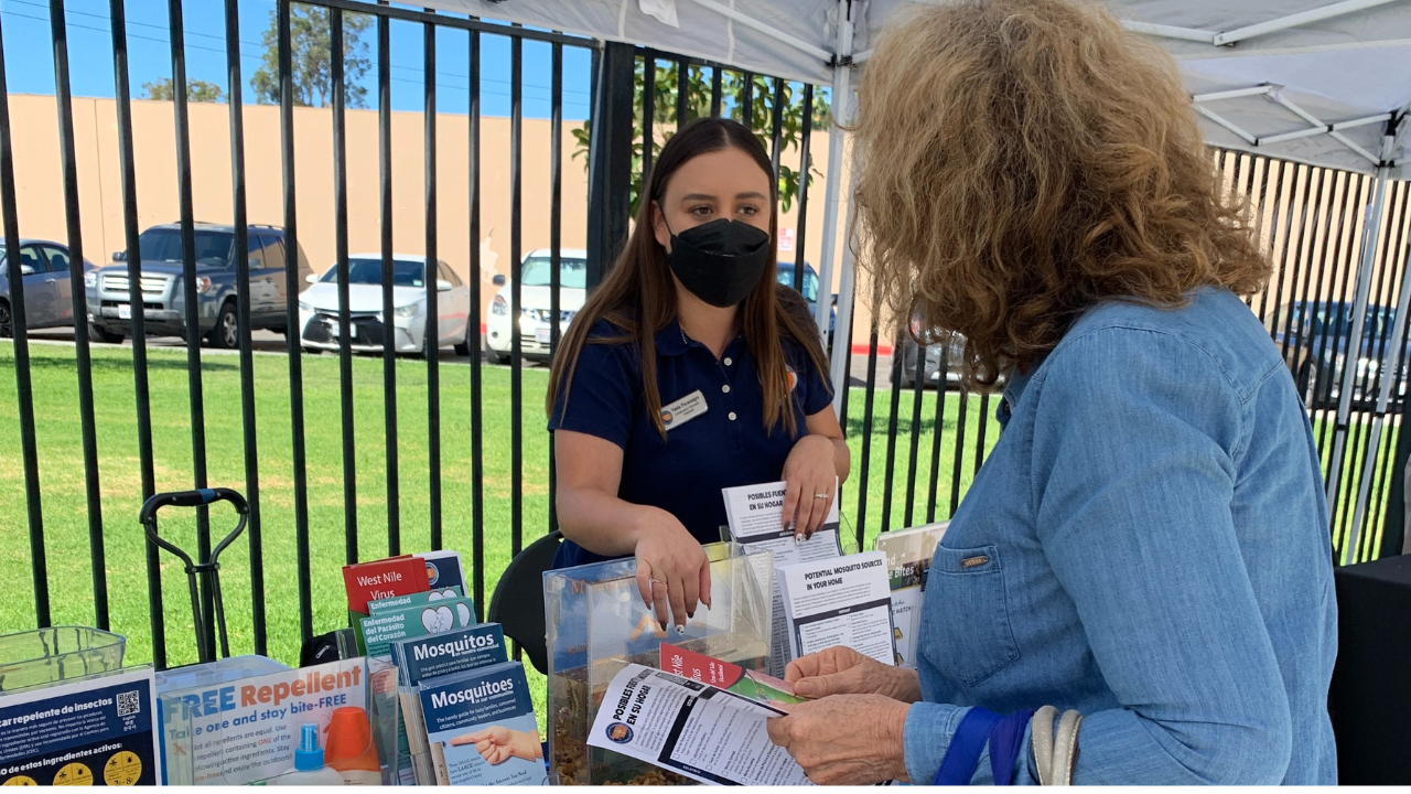 A woman at a booth talks with a representative about mosquito repellent and related informational materials.