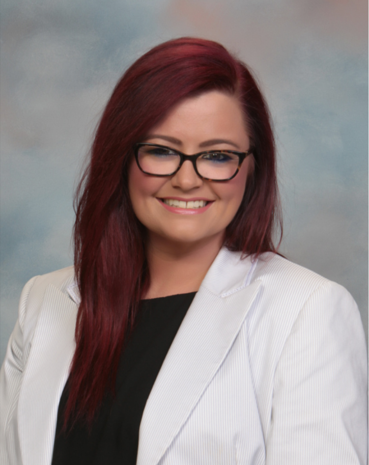A person with red hair and glasses smiling, wearing a white blazer and black top, against a cloudy background.