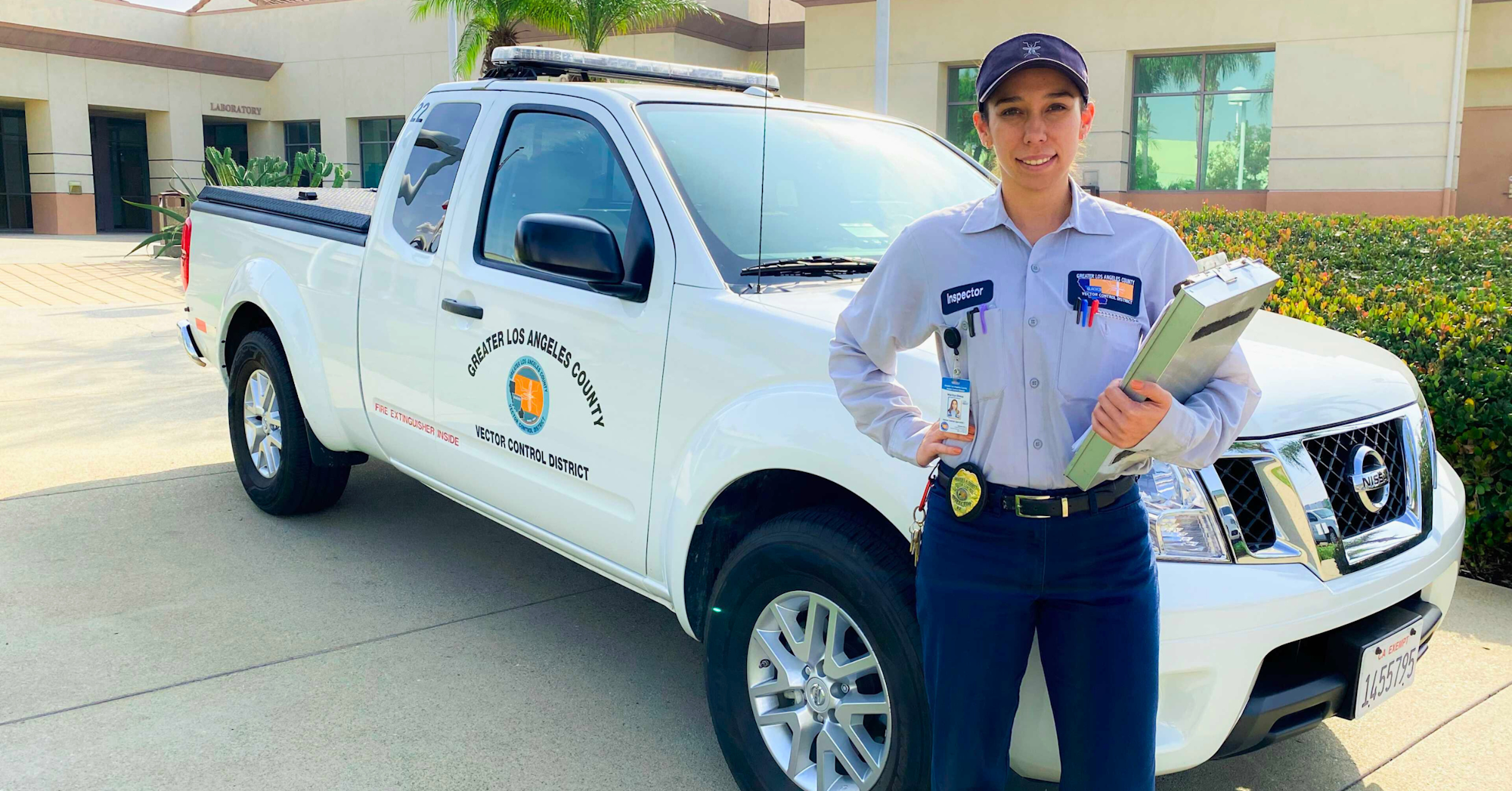 A smiling worker in uniform stands by a vector control district truck with a clipboard.
