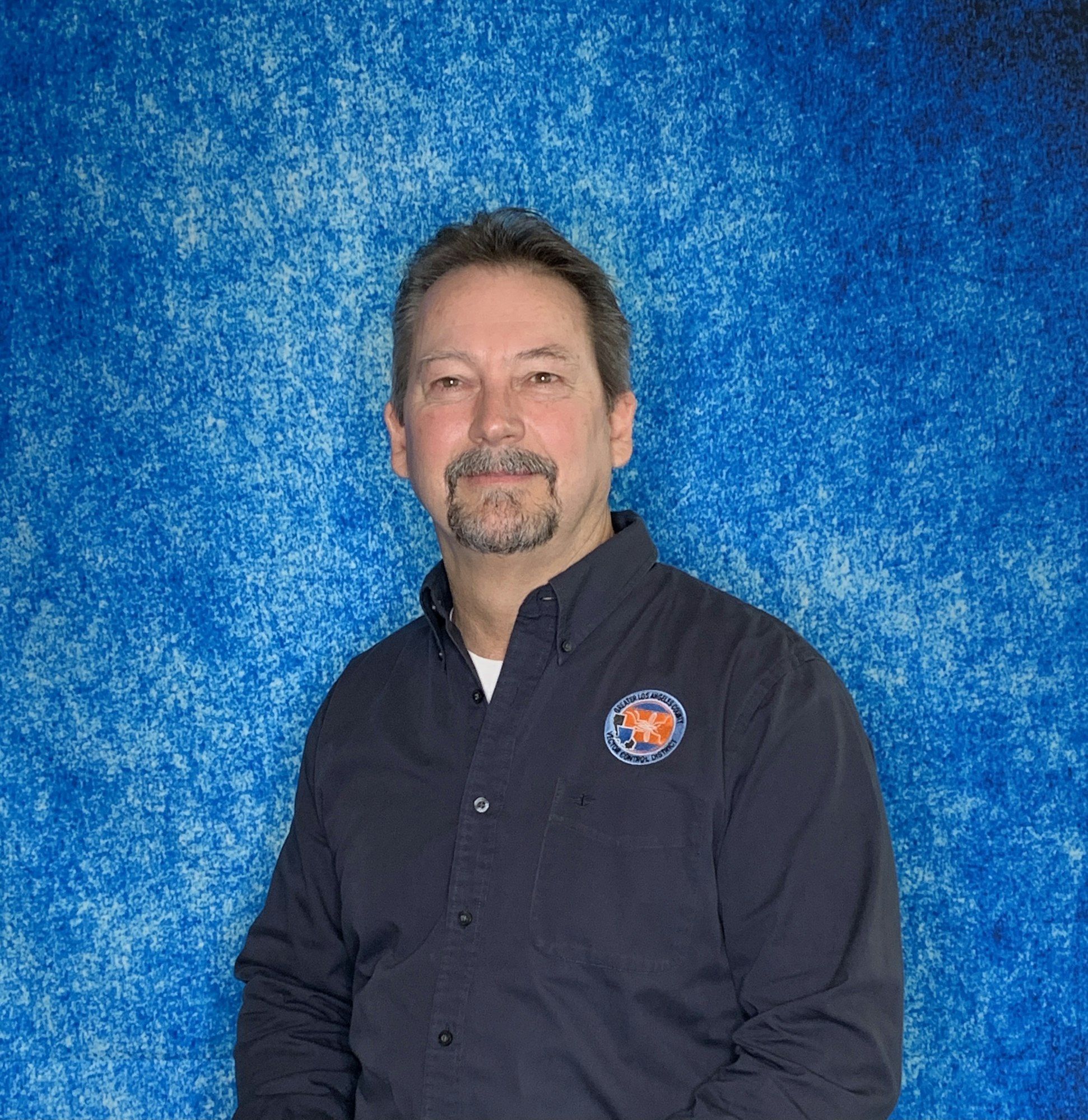 A man in a dark shirt with a logo stands against a blue textured background.