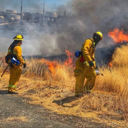 Firefighters battling a brush fire near industrial structures.