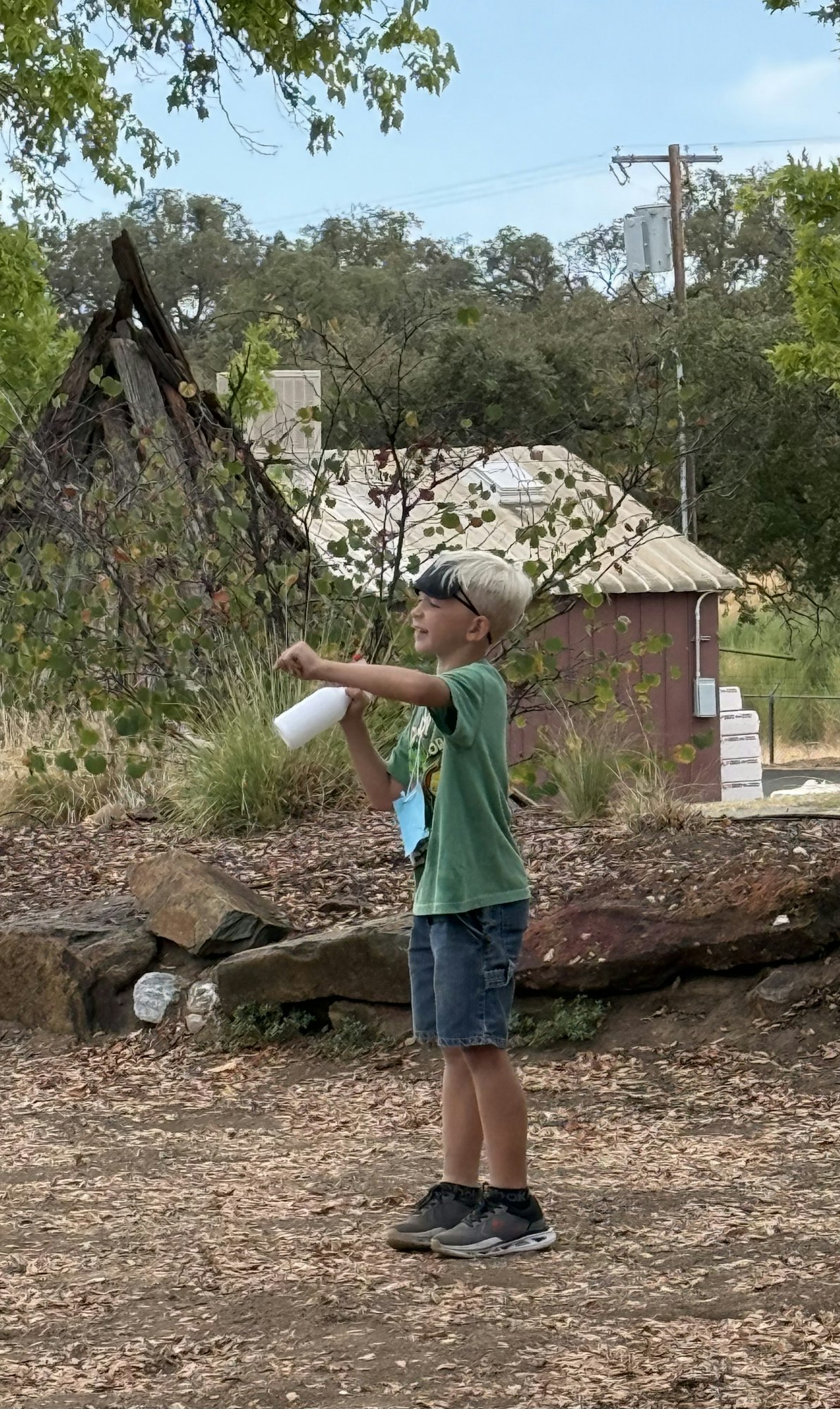 A child outdoors wearing a green shirt and blindfold, holding a bottle, surrounded by trees and a small building in the background.