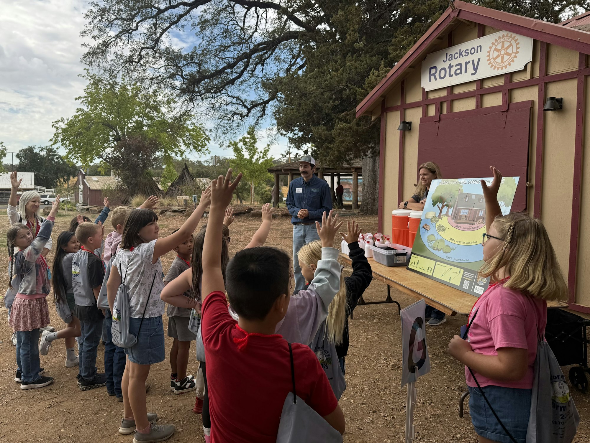 A group of children raising their hands outside at a "Jackson Rotary" booth with an educational poster, attended by two adults.