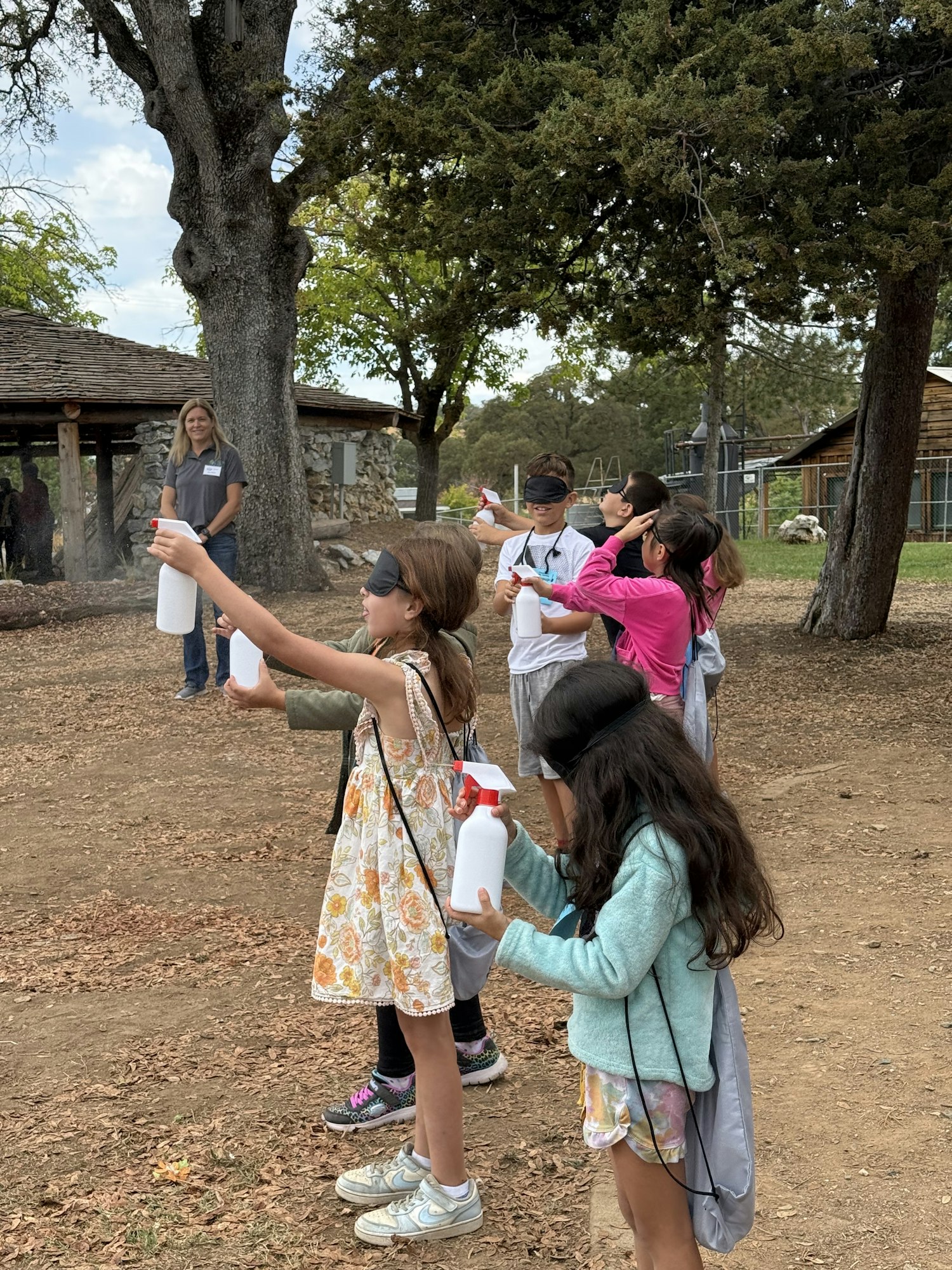 A group of children outdoors, holding spray bottles and wearing blindfolds in a line.