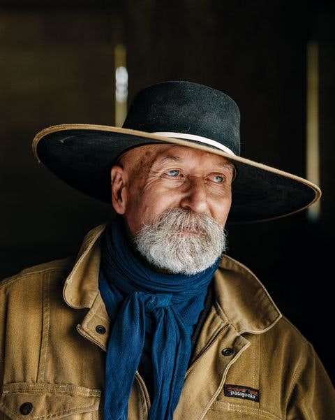 An older man with a beard, wearing a wide-brimmed hat and a blue scarf, gazes thoughtfully to the side.