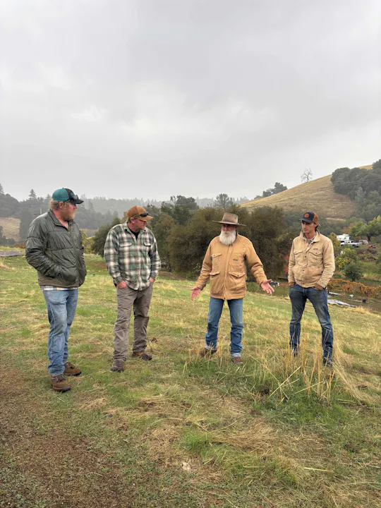 Four people in outdoor attire are discussing in a grassy area under cloudy skies, with a hilly backdrop.