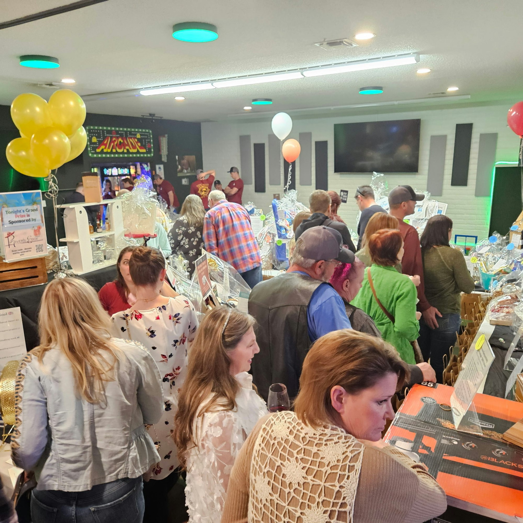 A crowded indoor event with people browsing tables, balloons, and arcade machines in the background.