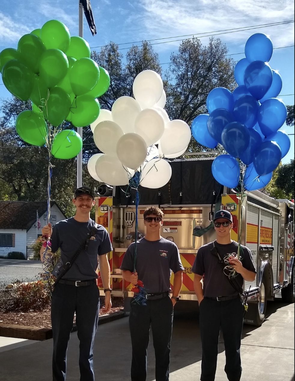 Three people holding green, white, and blue balloons in front of a fire truck.