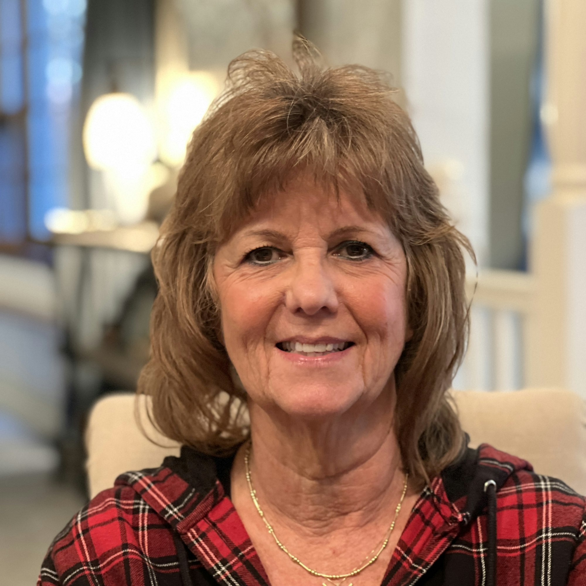 A person smiling, wearing a red plaid top, indoors with blurred background lighting.