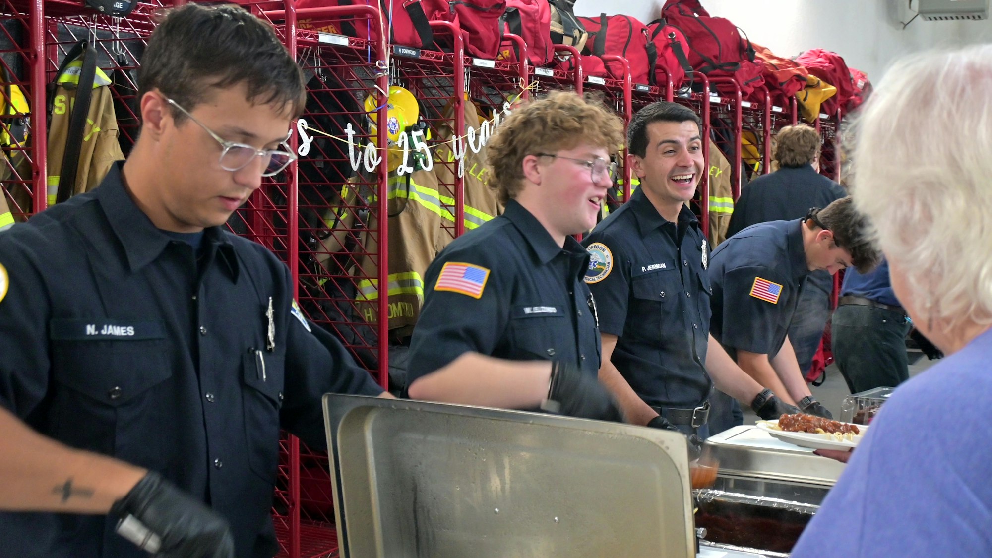 Firefighters in uniform serve food in a station with gear in the background, interacting with an elderly person.