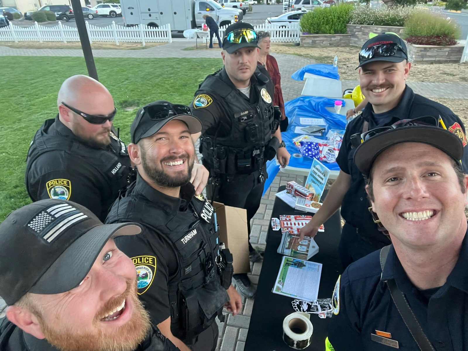 Group of officers and a firefighter smiling at an outdoor event table with brochures and items.