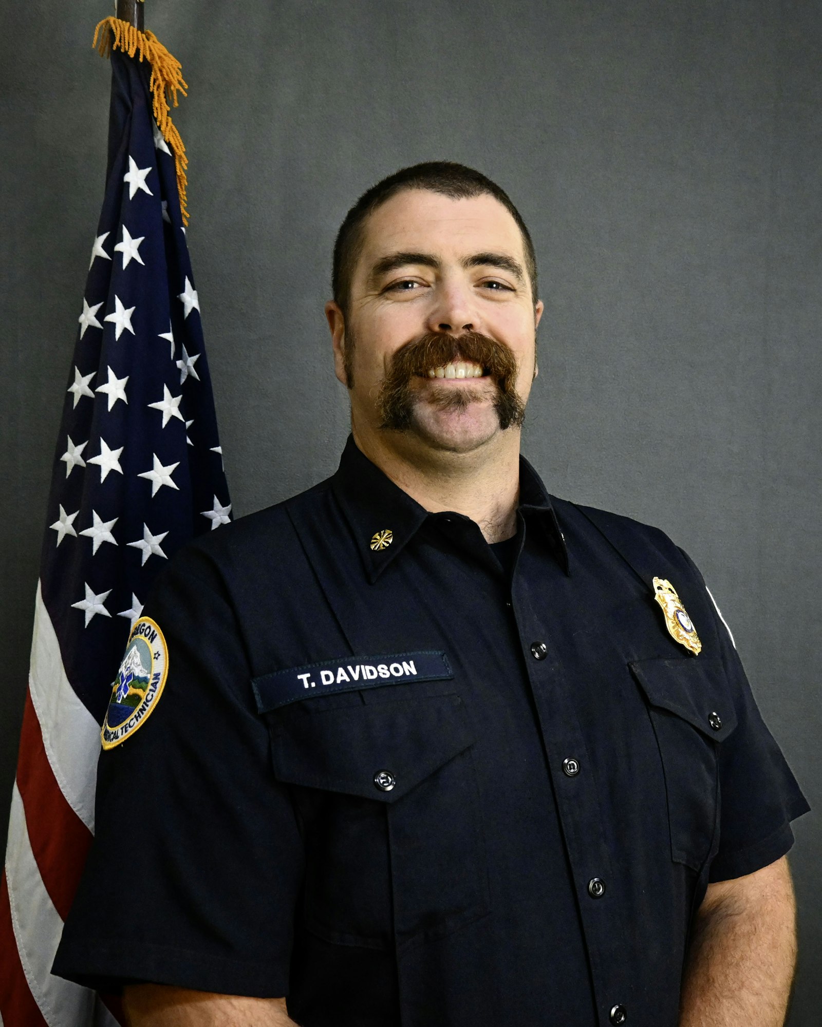 A man in uniform stands smiling next to an American flag.