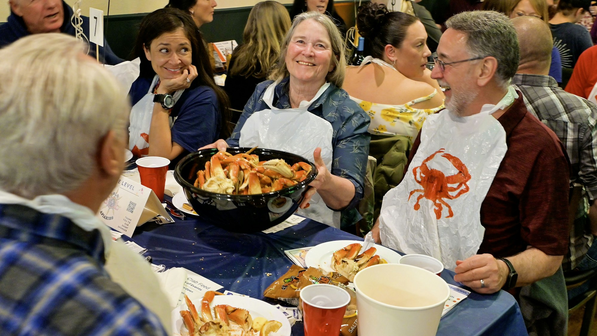 People enjoying a crab feast, wearing bibs, and sharing a large bowl of crabs at a table.
