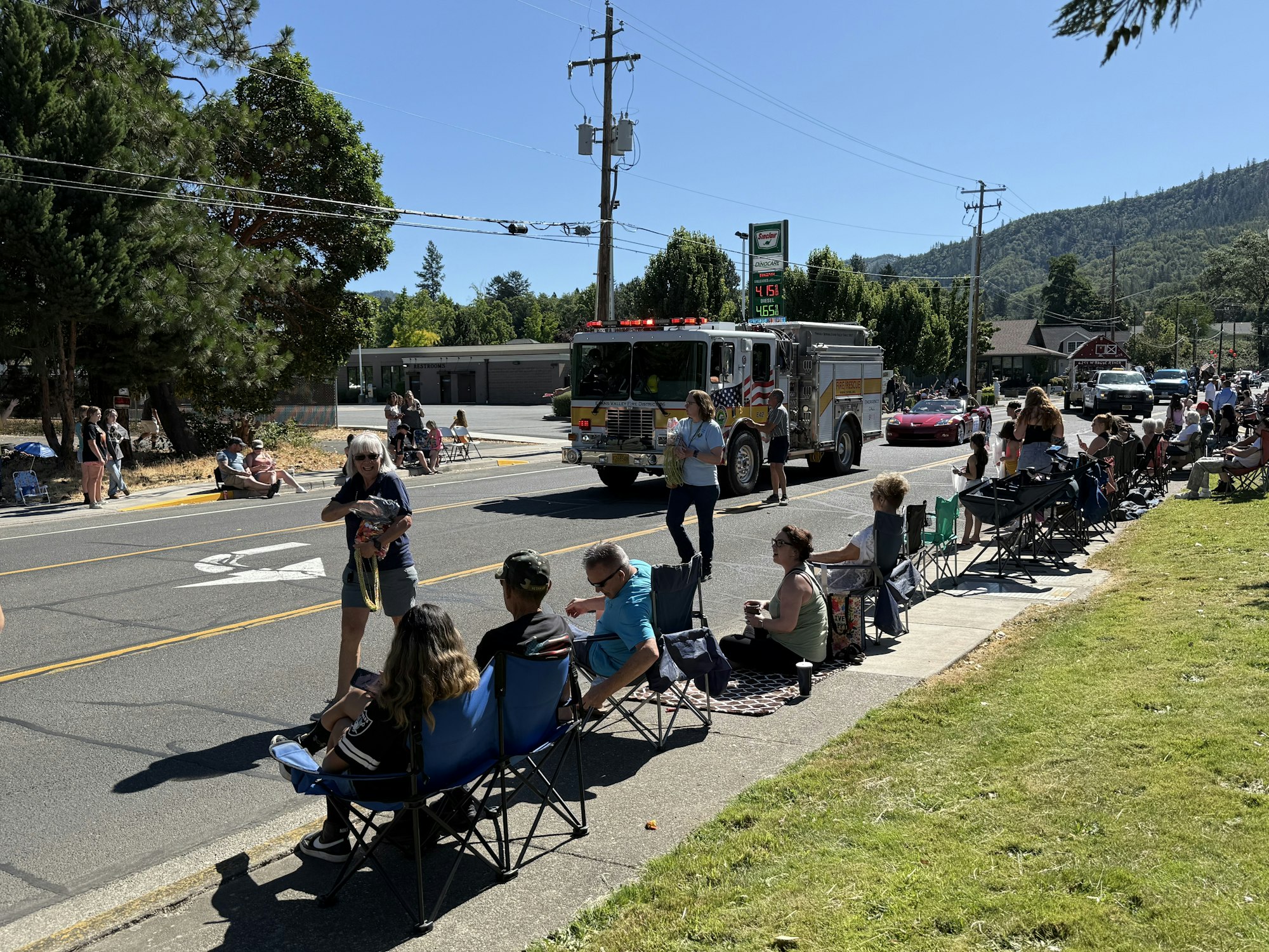 People watch a parade with a fire truck passing by; others are sitting on chairs alongside the street.