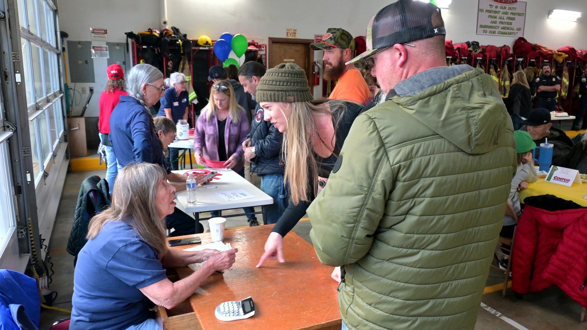 People interacting at a community event inside a room with tables, balloons, and safety gear on racks.