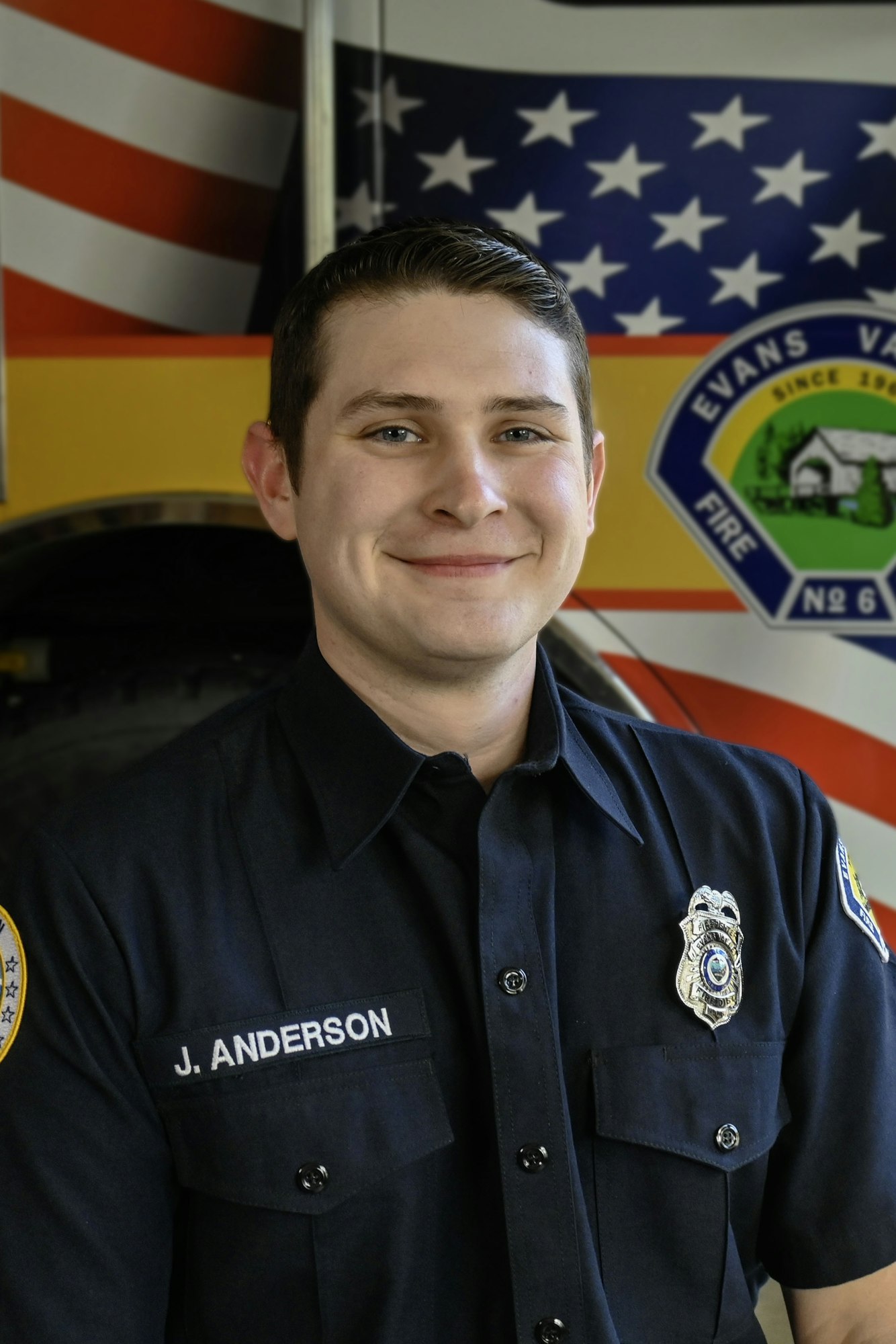 A smiling individual in a fire department uniform stands in front of a truck displaying an American flag and insignia.