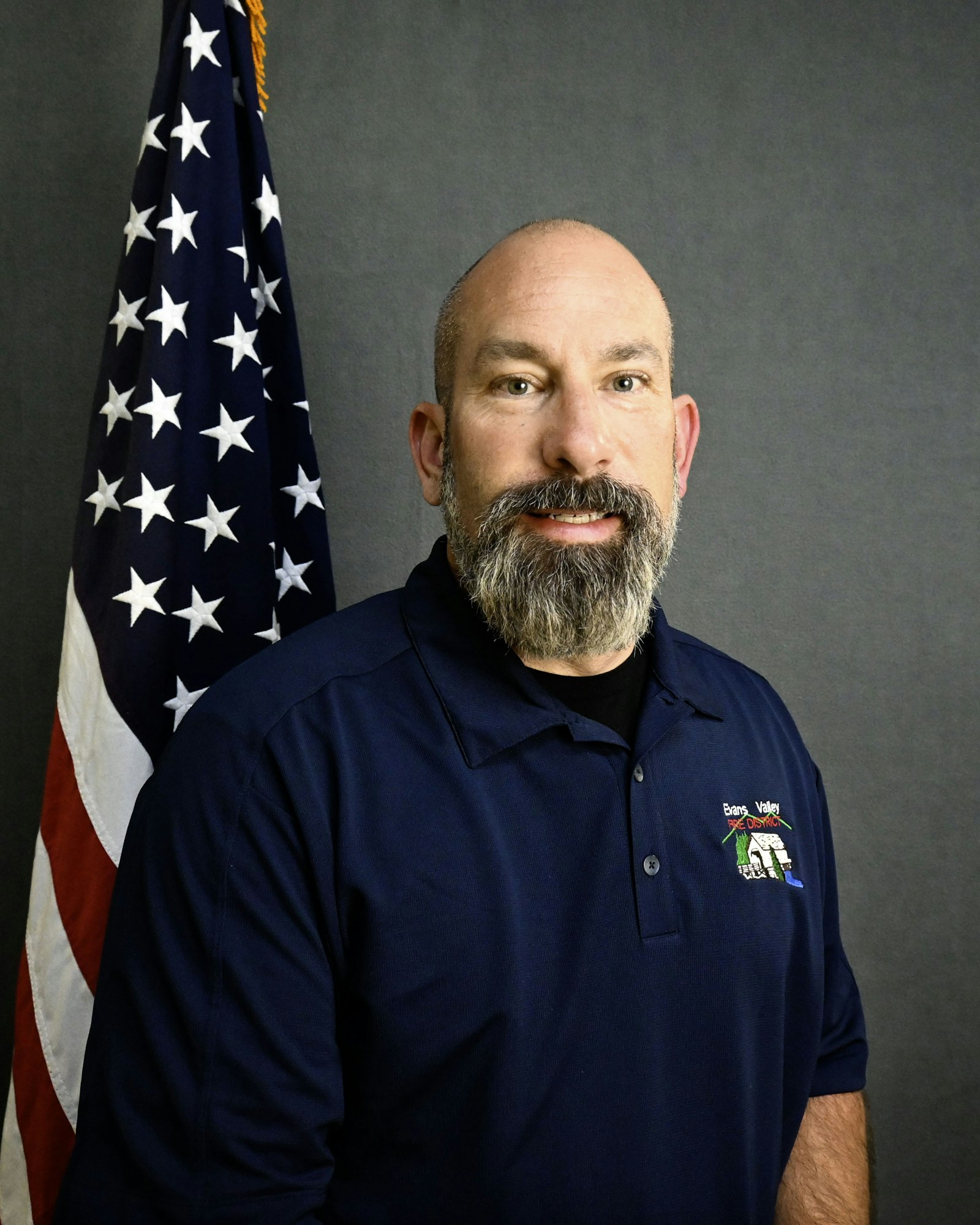A man with a beard in a blue shirt stands next to an American flag.