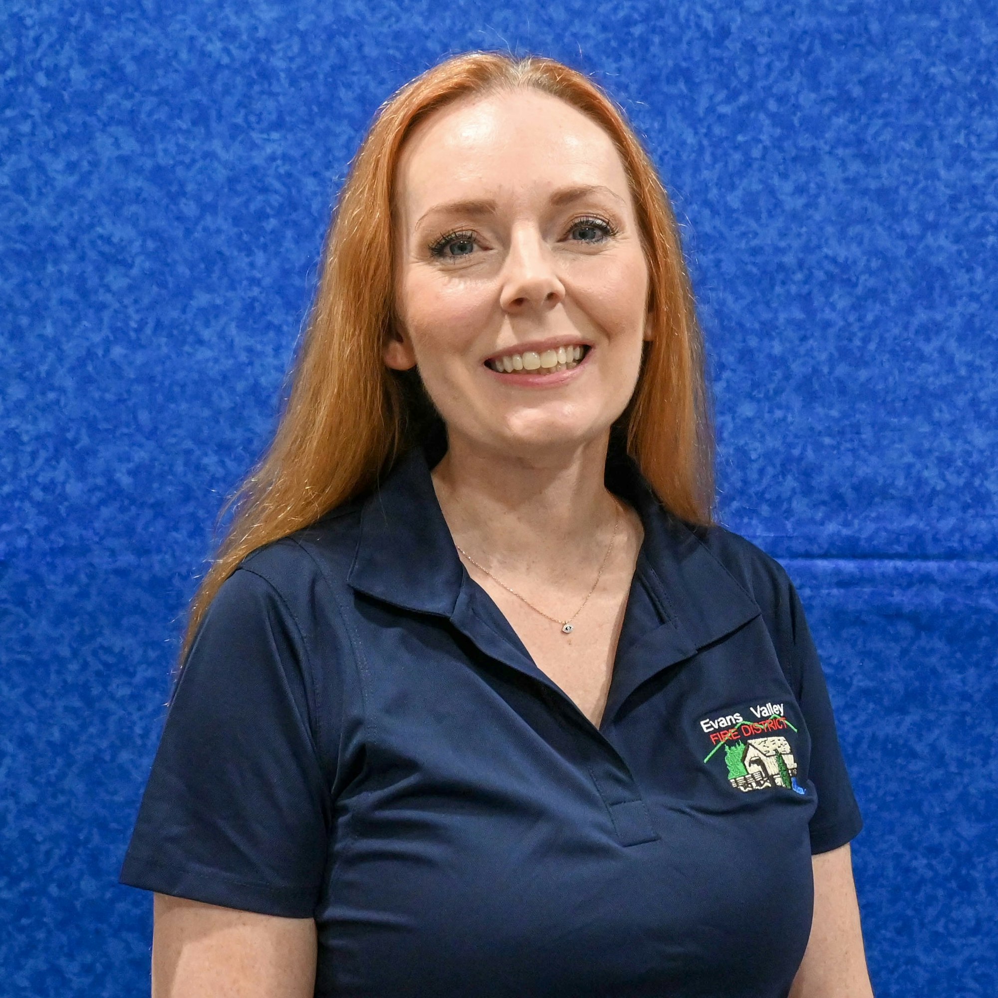 A smiling person with long hair is wearing a navy polo shirt against a blue background.