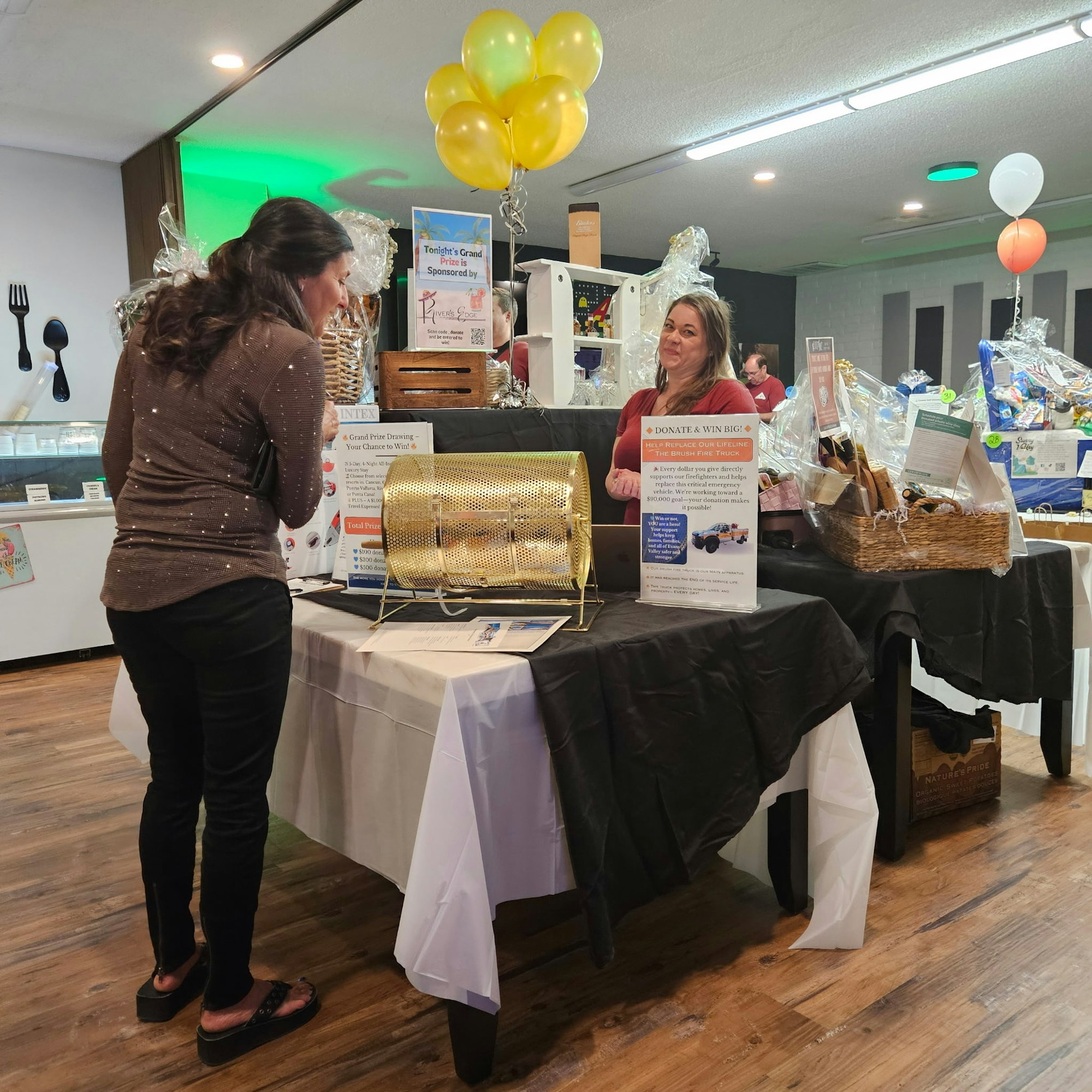 Two people at a raffle table with gift baskets and balloons.