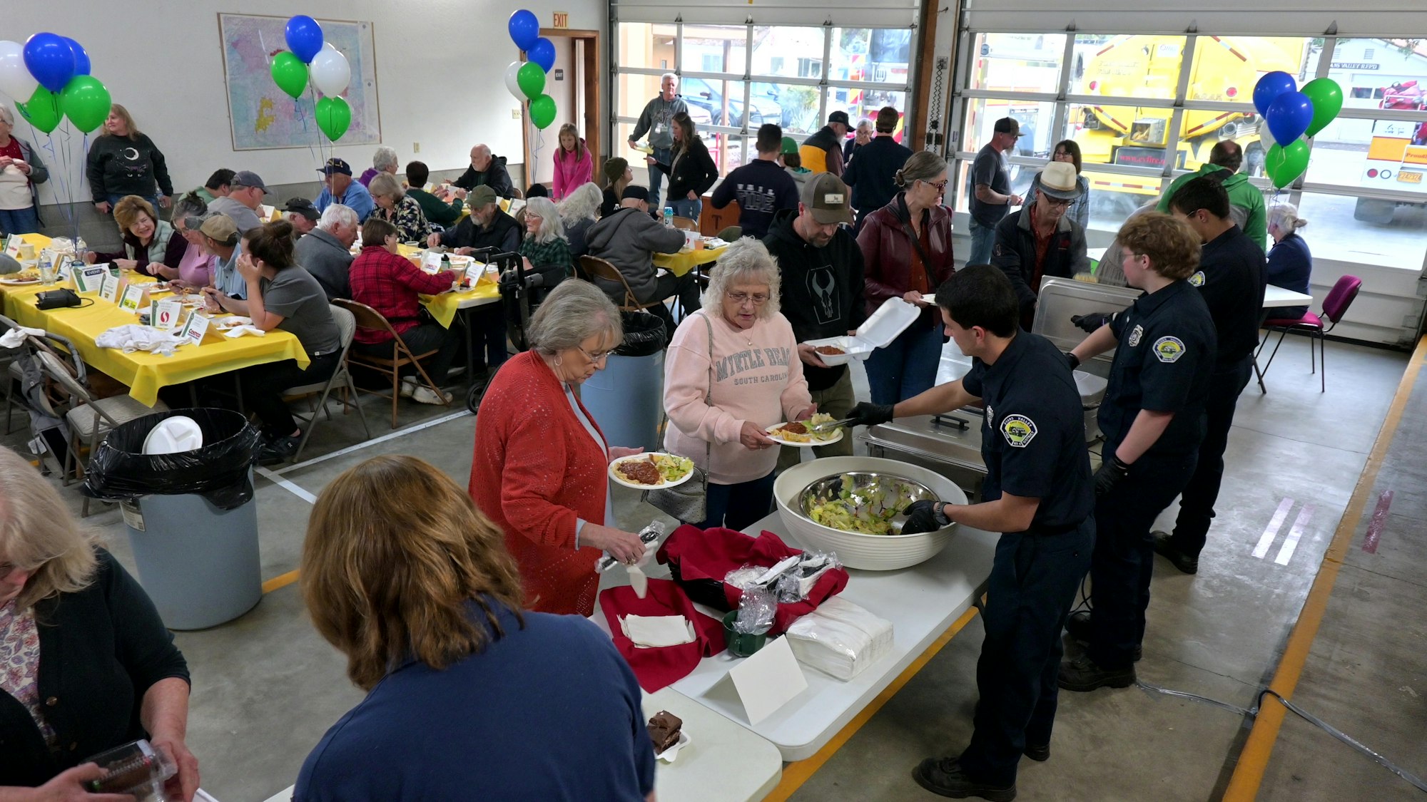 Community gathering with people having a meal; servers in uniform; tables with decorations and balloons.
