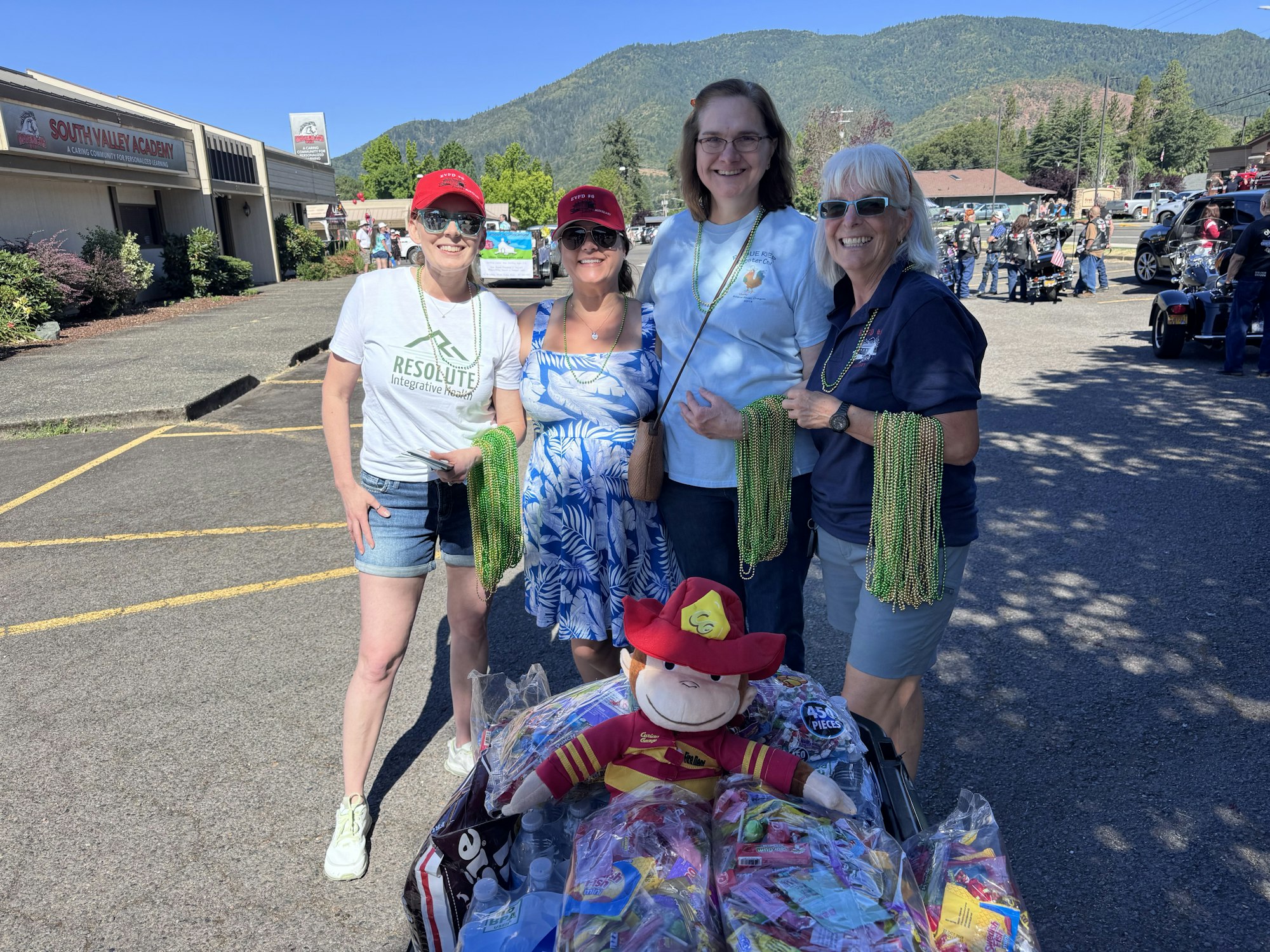 Four people holding green bead necklaces and a stuffed animal, standing beside a table with colorful bags and items outdoors.