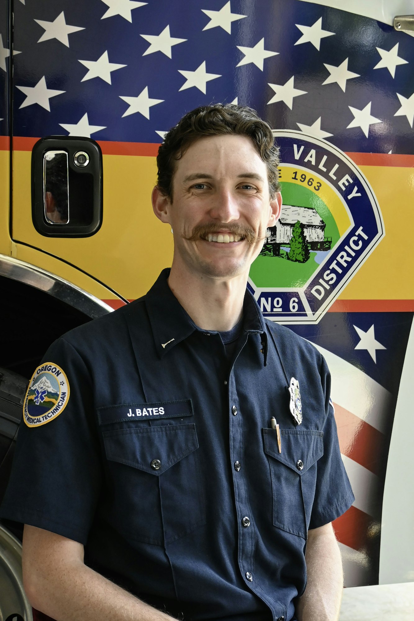 A person in a uniform with "J. Bates" on the badge, standing in front of a fire district vehicle with a star-spangled design.