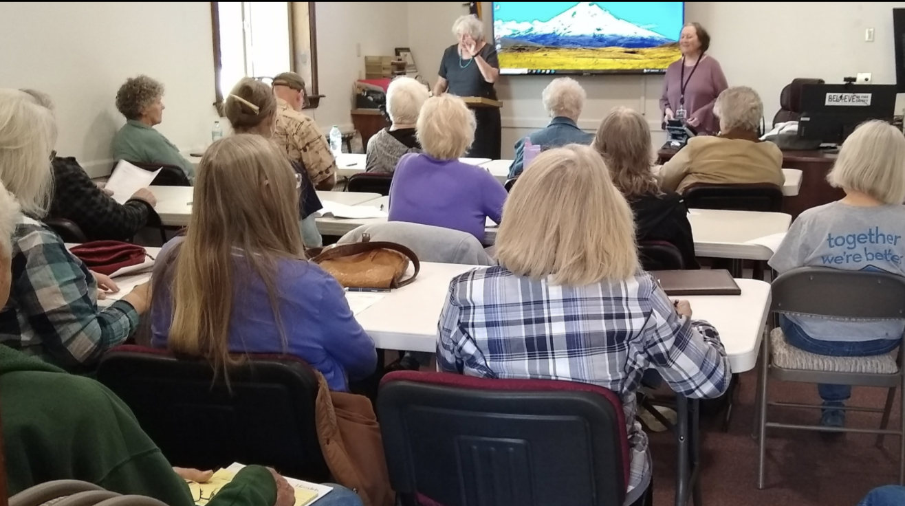 A group of people seated in a classroom setting, listening to a speaker at the front, with a screen displaying a mountain landscape.