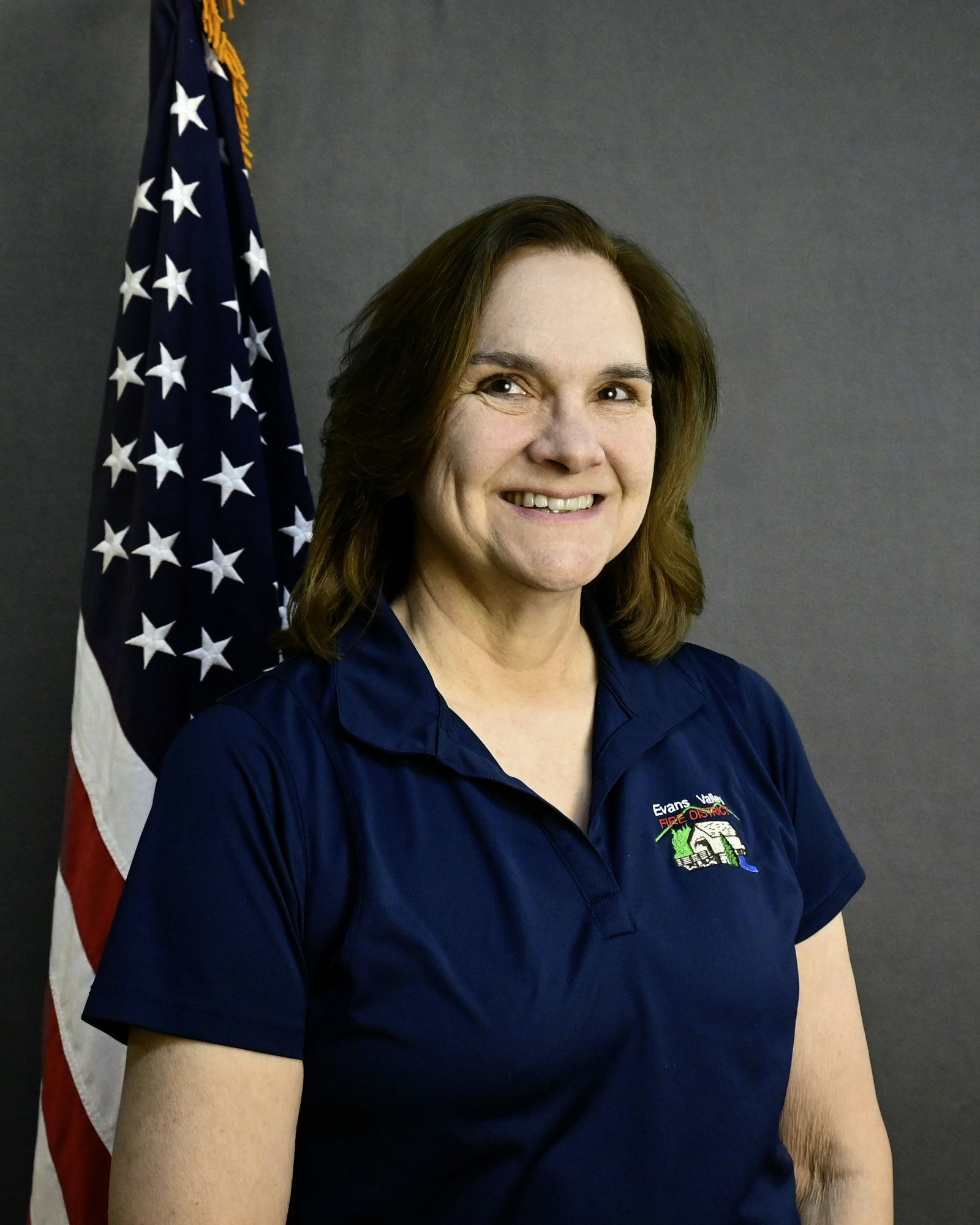 A person wearing a navy polo shirt stands beside a U.S. flag, smiling against a plain background.