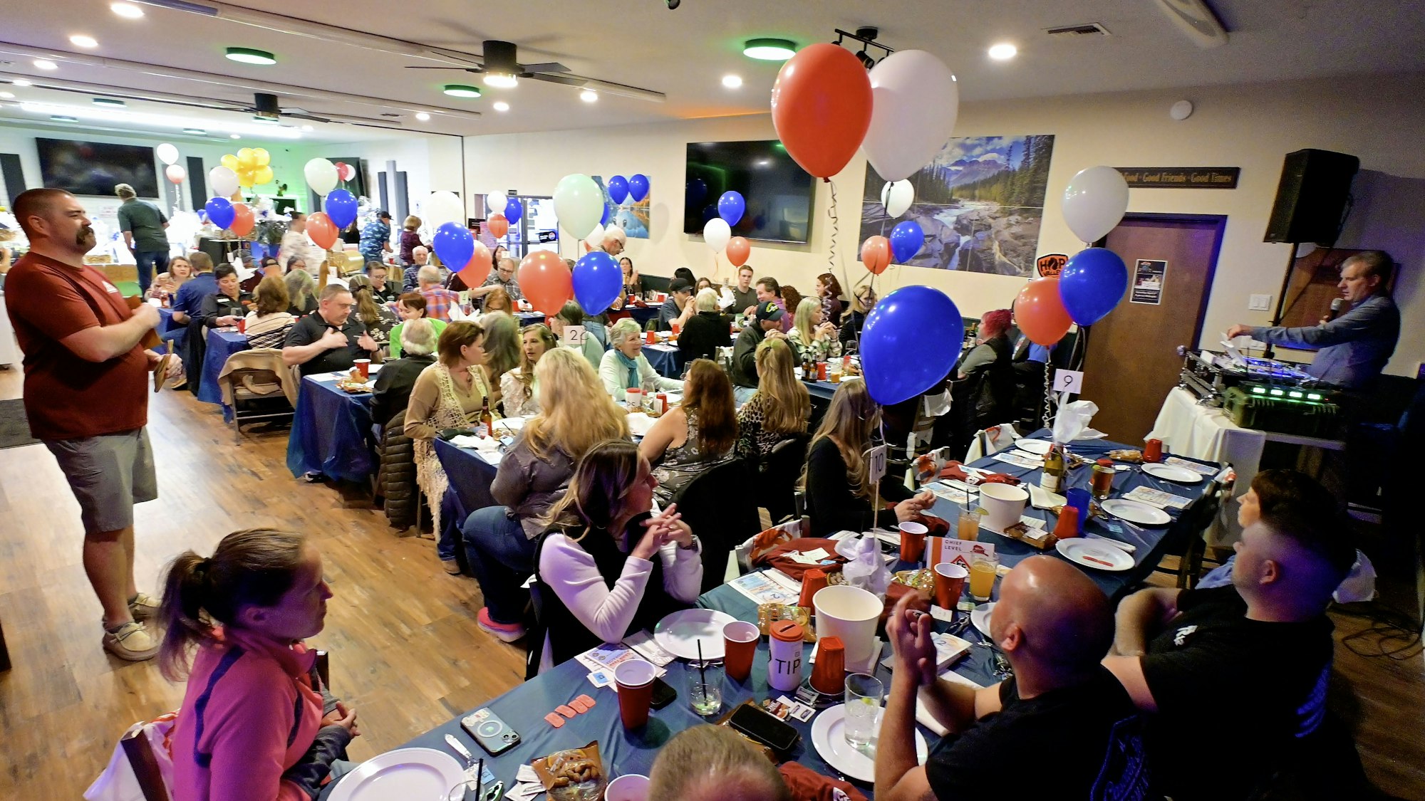 A lively indoor event with balloons and people seated at tables, listening to a speaker.