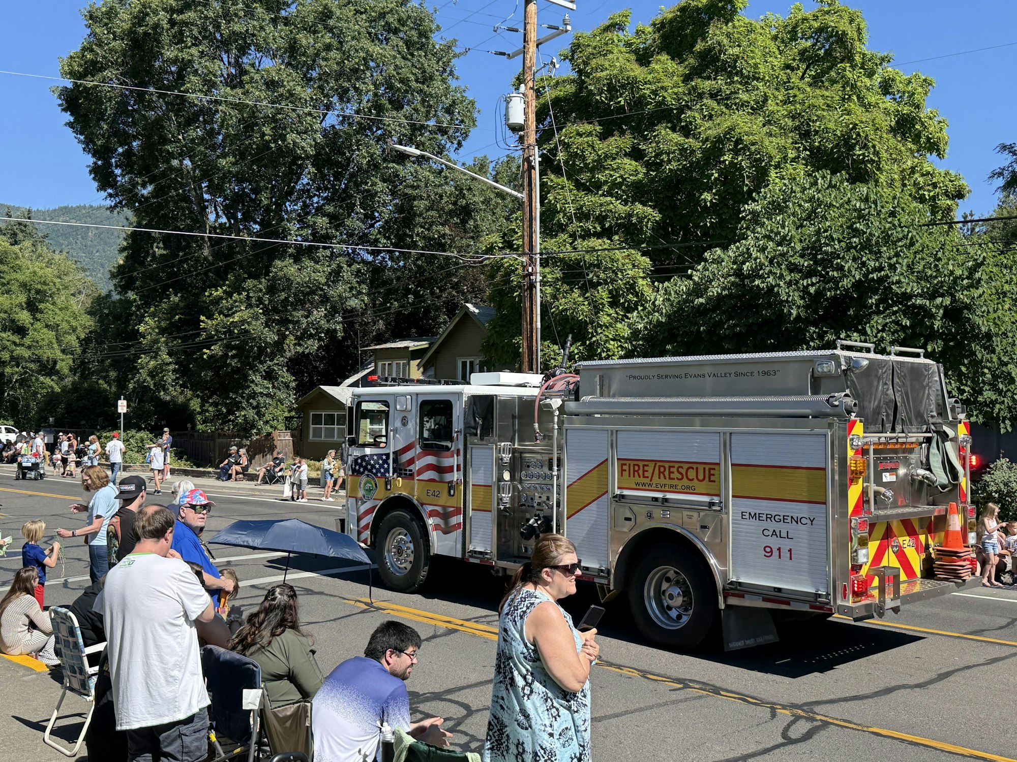 A fire truck in a street parade, with people sitting and standing on the sidewalk, some with umbrellas.