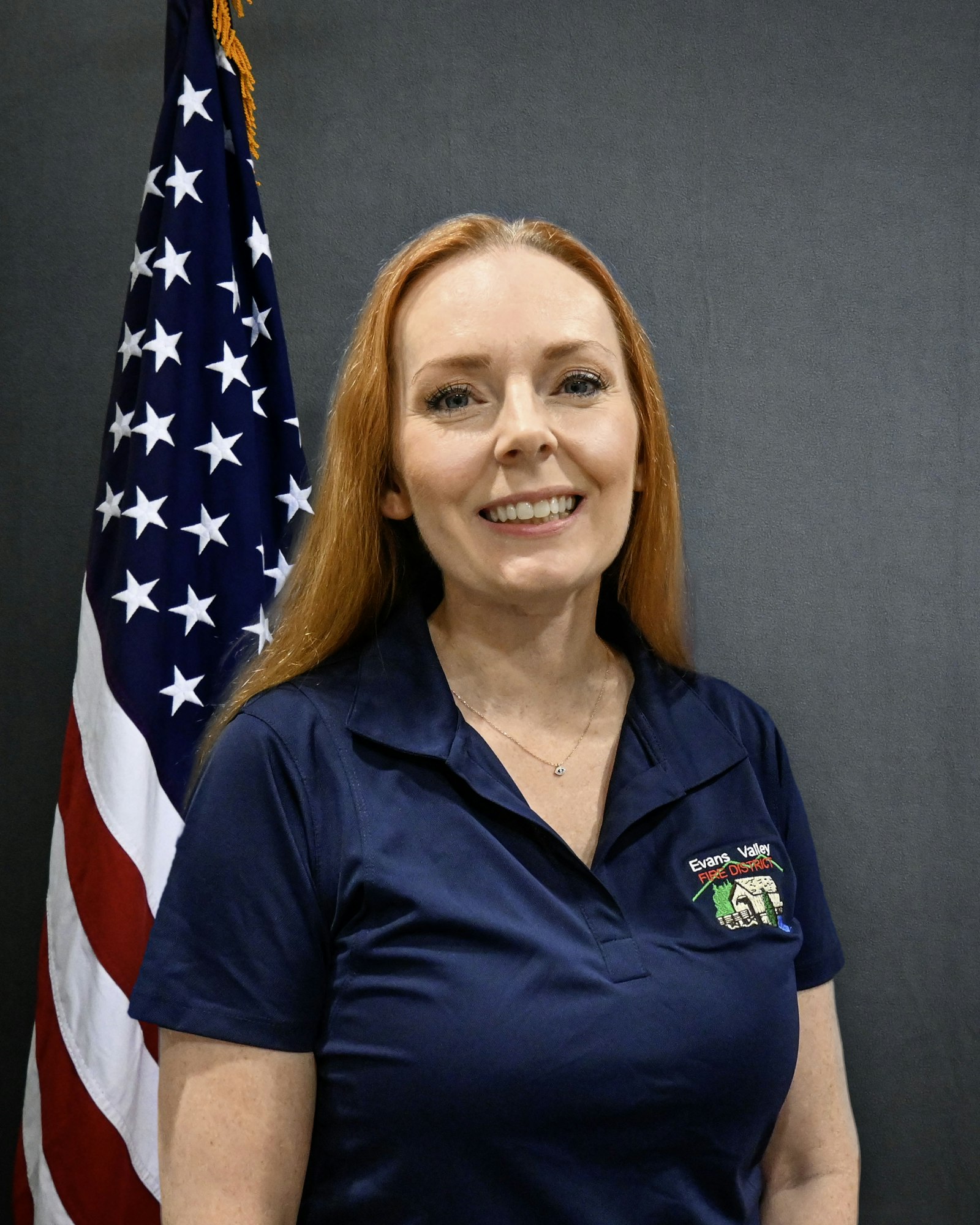 A person with red hair in a navy shirt stands in front of the U.S. flag with a gray background.