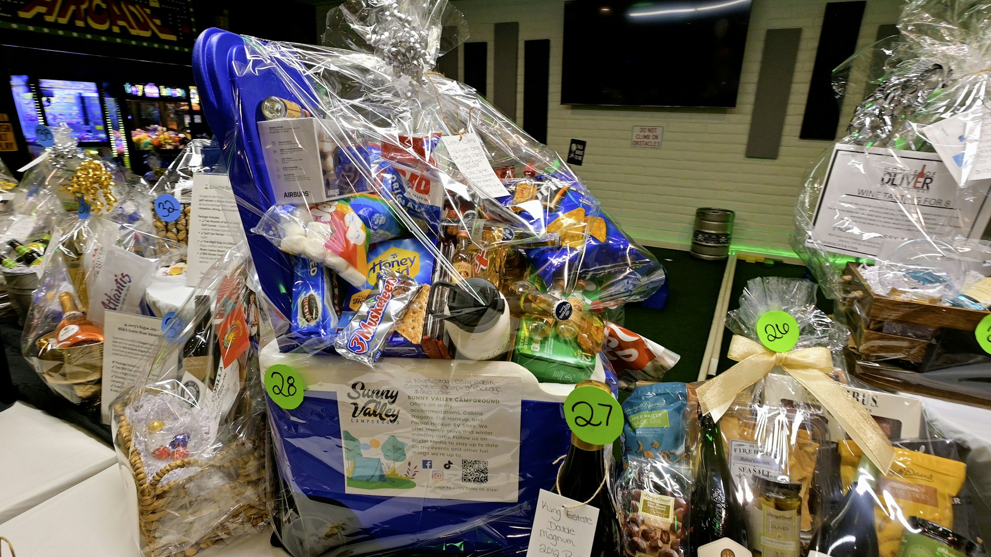 Gift baskets with snacks, drinks, and goodies wrapped in cellophane, labeled with numbers. Indoor setting with arcade games in the background.