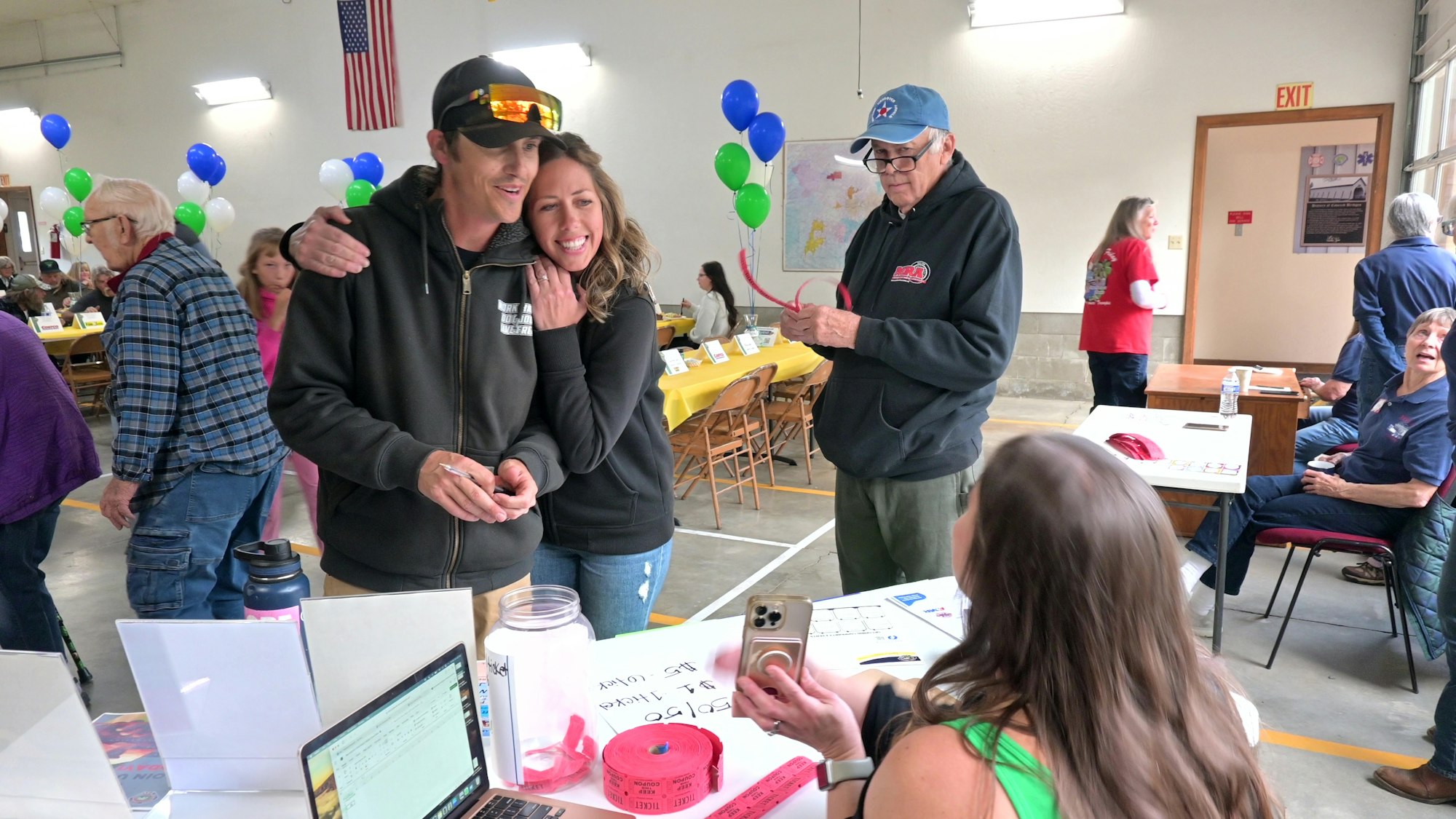 People at an indoor event with tables, balloons, and ticket purchases happening.