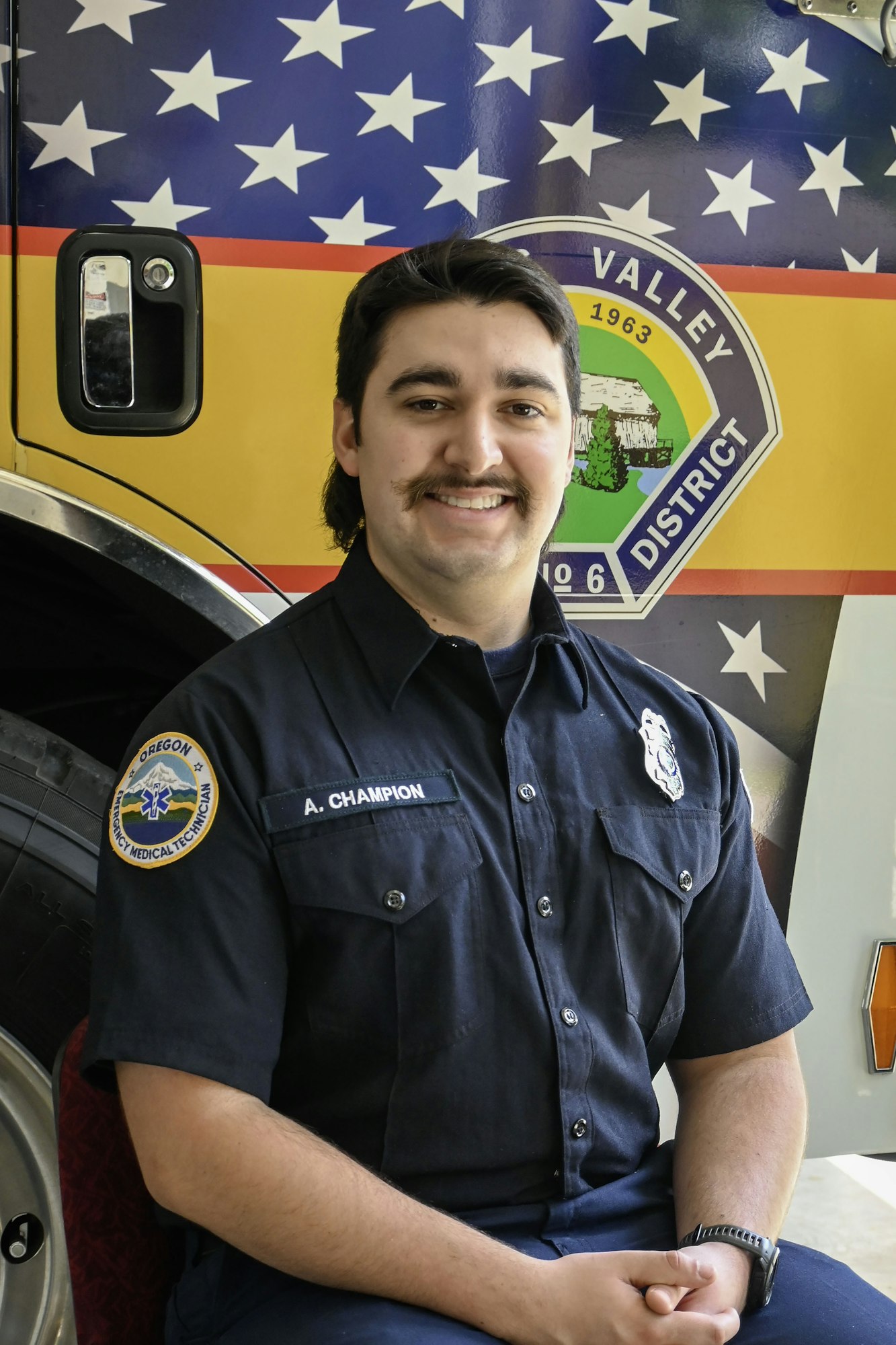 A firefighter sits smiling, wearing a uniform with a patch. There's an American-themed fire truck in the background.