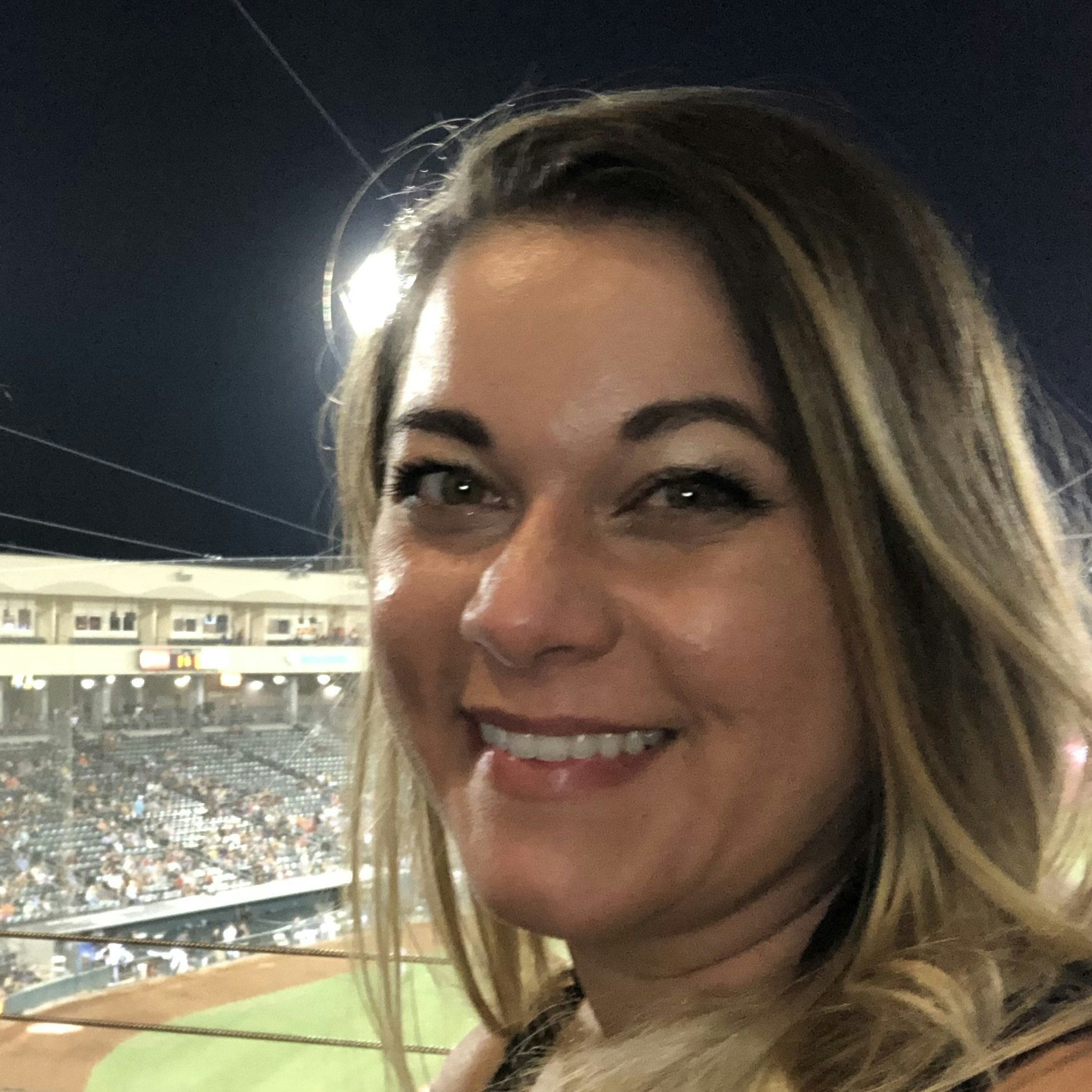 A person smiling at a night-time sports event, likely a baseball game, with the field and spectators in the background.