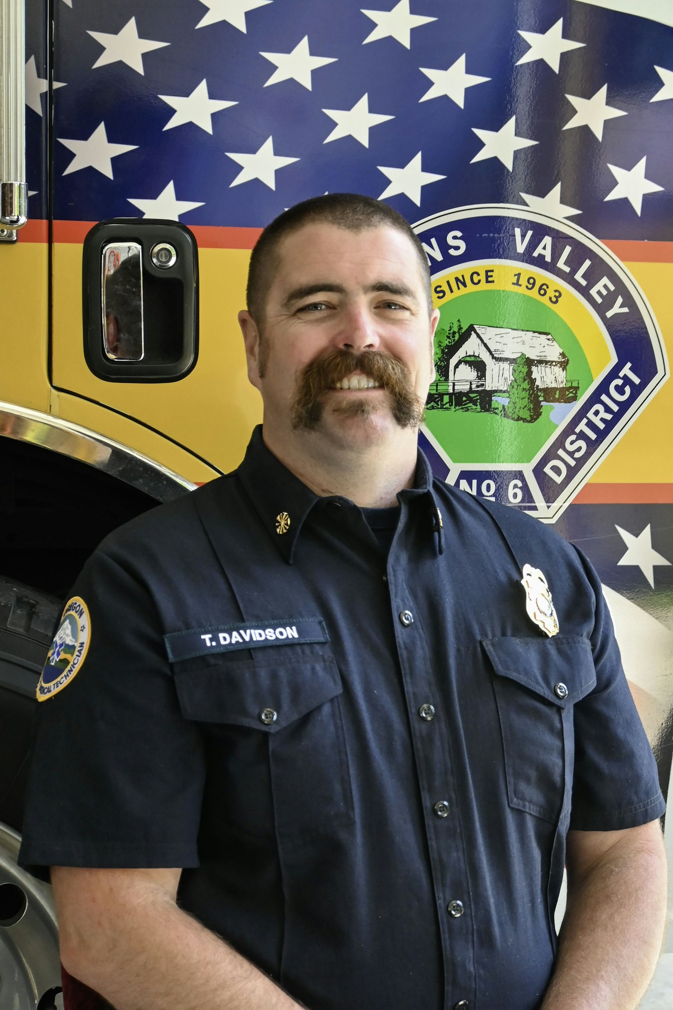 A firefighter in uniform stands in front of a fire truck with a flag design.