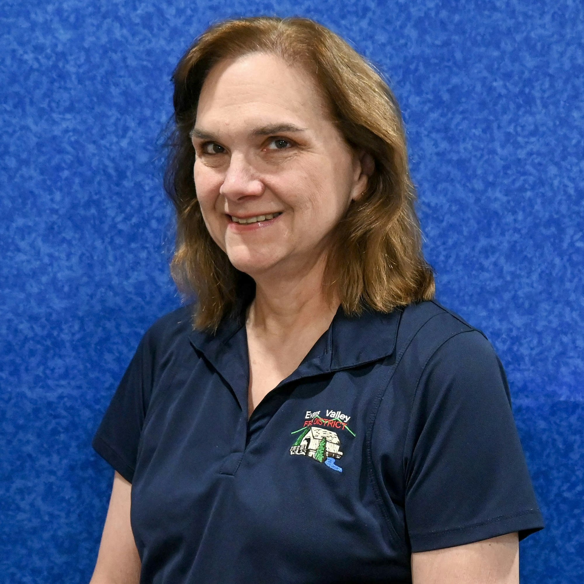 A person wearing a dark blue polo shirt with a logo on it, standing against a blue background.