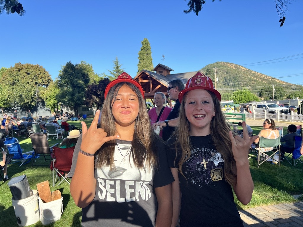 Two people outdoors wearing red firefighter helmets, smiling and making hand signs.