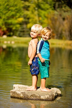 Two children standing on a rock in a pond, smiling and wearing swimwear.