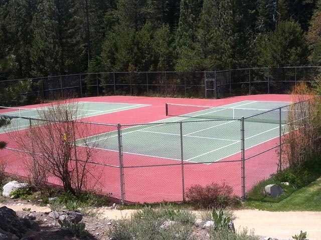 A tennis court surrounded by a fence, located in a wooded area with trees in the background.
