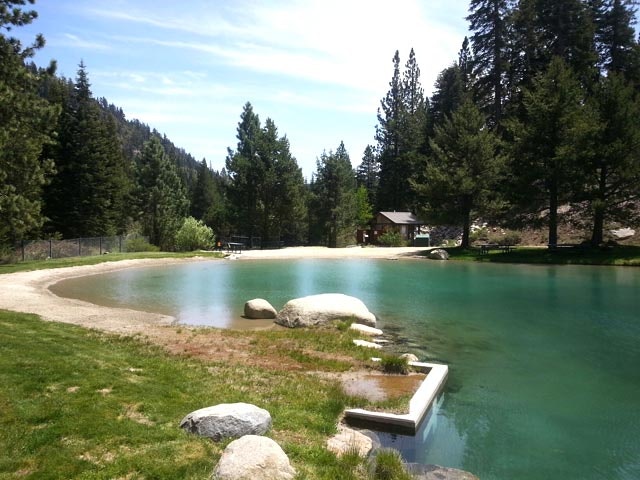 A serene pond surrounded by rocks and trees, with a small building in the background under a clear blue sky.