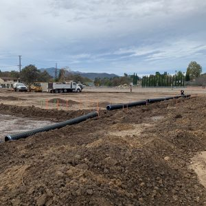 Construction site with large pipes laid in trenches and trucks in the background, under a cloudy sky.