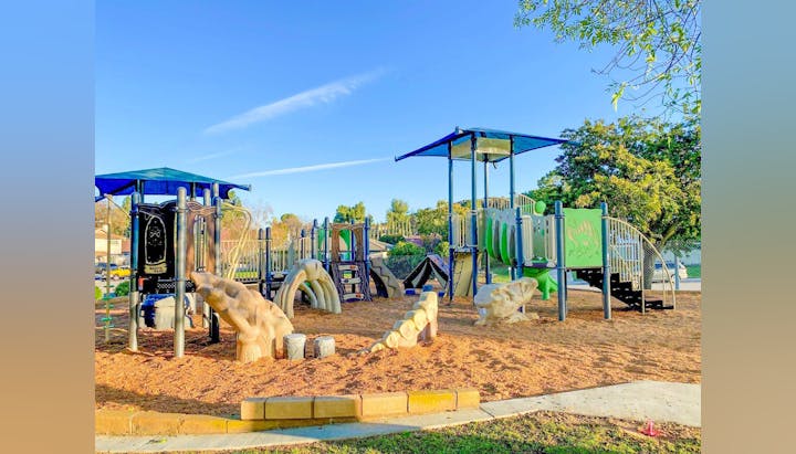 A colorful playground with slides, climbing structures, and wood chips on the ground, set in a park with trees and a clear sky.