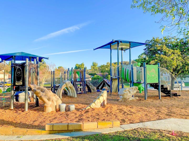 A colorful playground with slides, climbing structures, and wood chips on the ground, set in a park with trees and a clear sky.